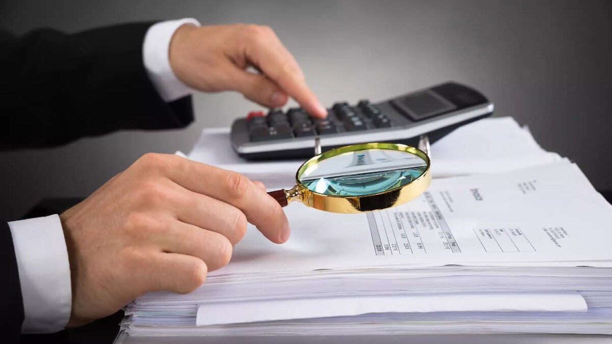 Close-up Of Businessperson Hands Checking Invoice With Magnifying Glass At Desk