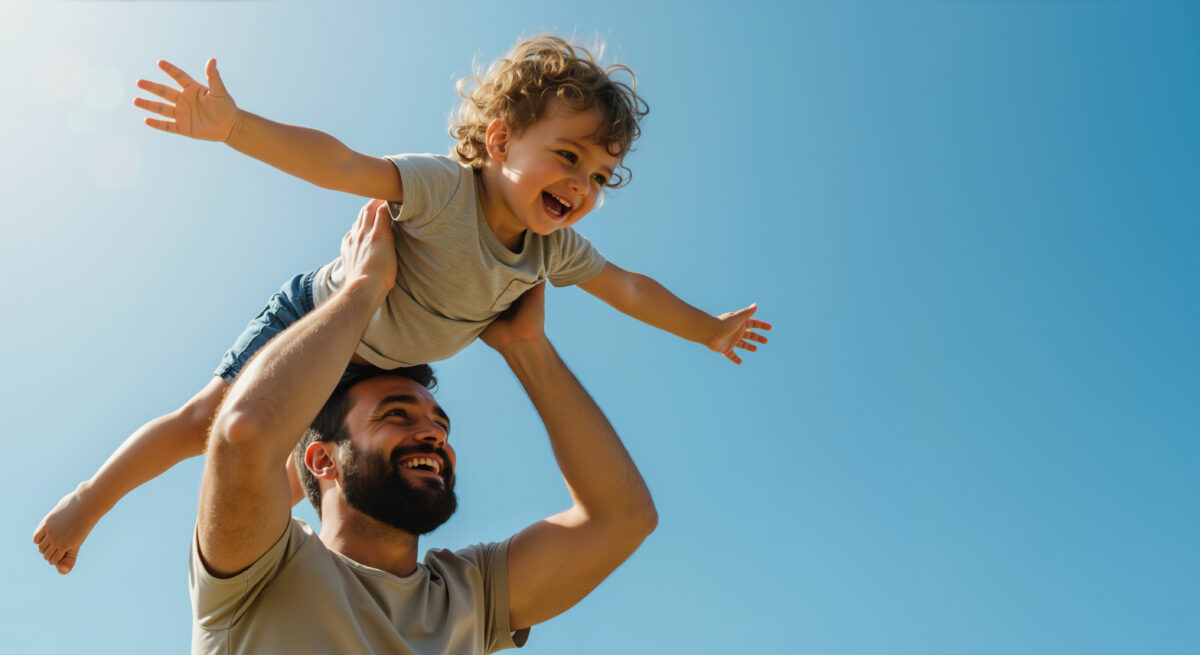 Happy bearded father lifting curly haired toddler son with arms spread against blue sky. Father's day celebration and family bonding activities