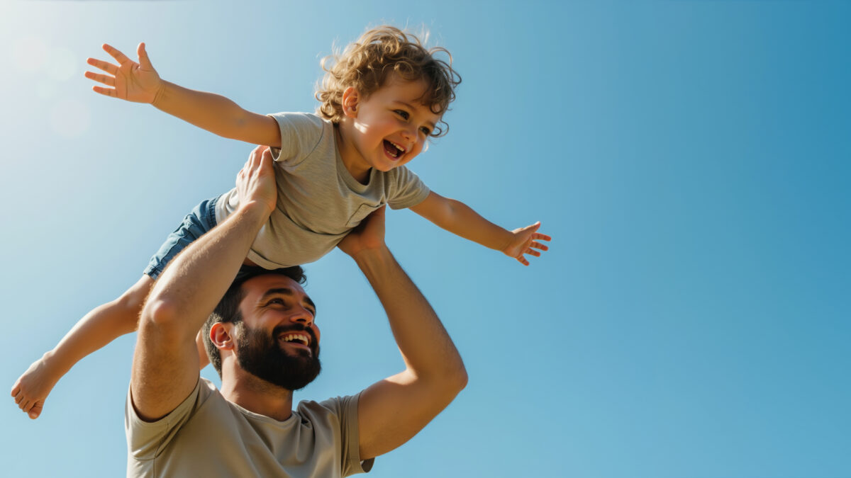 Happy bearded father lifting curly haired toddler son with arms spread against blue sky. Father's day celebration and family bonding activities