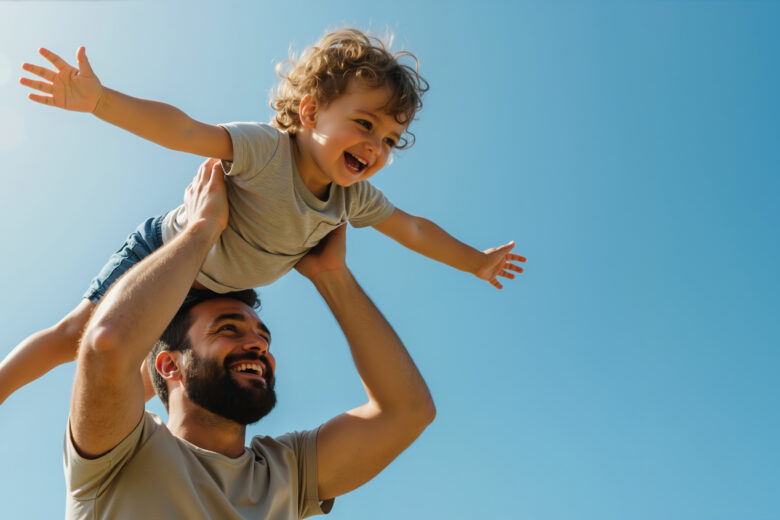 Happy bearded father lifting curly haired toddler son with arms spread against blue sky. Father's day celebration and family bonding activities