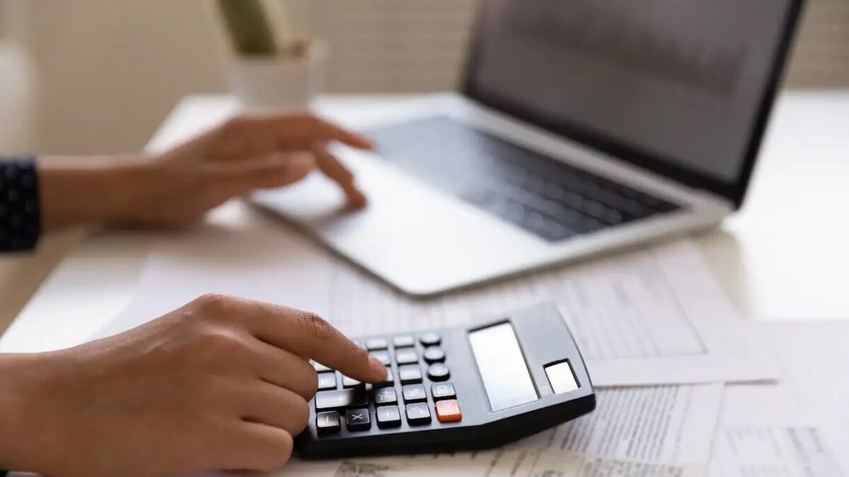 Close up of young female electronic bank client using calculator in paperwork with accounts bills before providing payment online by laptop. Woman hands at work table with papers accounting sum to pay