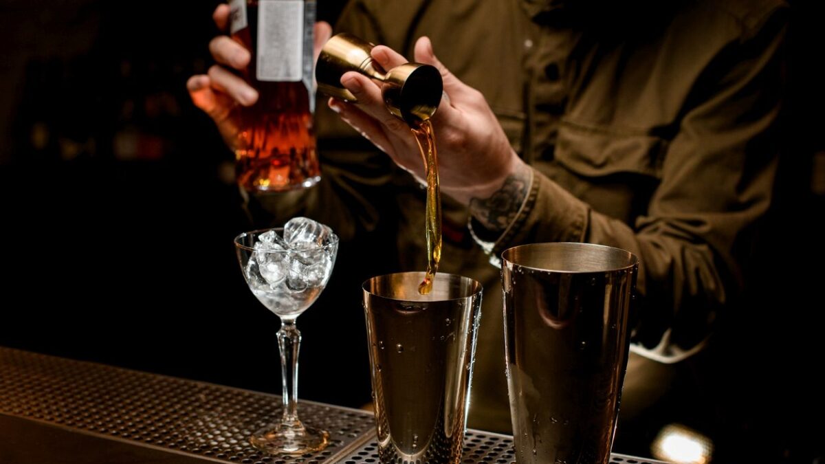 male bartender pours alcoholic drink from jigger into cup of steel cocktail shaker. Glass of ice is on bar