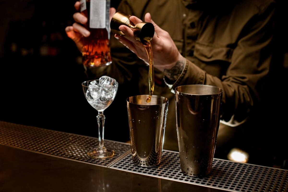 male bartender pours alcoholic drink from jigger into cup of steel cocktail shaker. Glass of ice is on bar