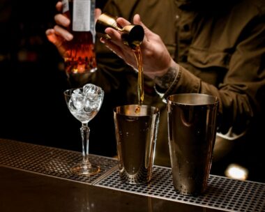 male bartender pours alcoholic drink from jigger into cup of steel cocktail shaker. Glass of ice is on bar