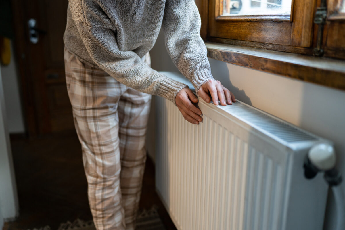 Closeup of woman warming her hands on the heater at home during cold winter days, top view. Female in woolen sweater getting warm up her arms over radiator. Concept of heating season, cold weather.