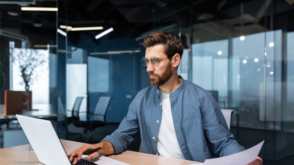 A serious young man accountant, financier, analyst, auditor sits in the office at the table. He holds documents and a pen in his hands, checks accounts, finances, types on a laptop.