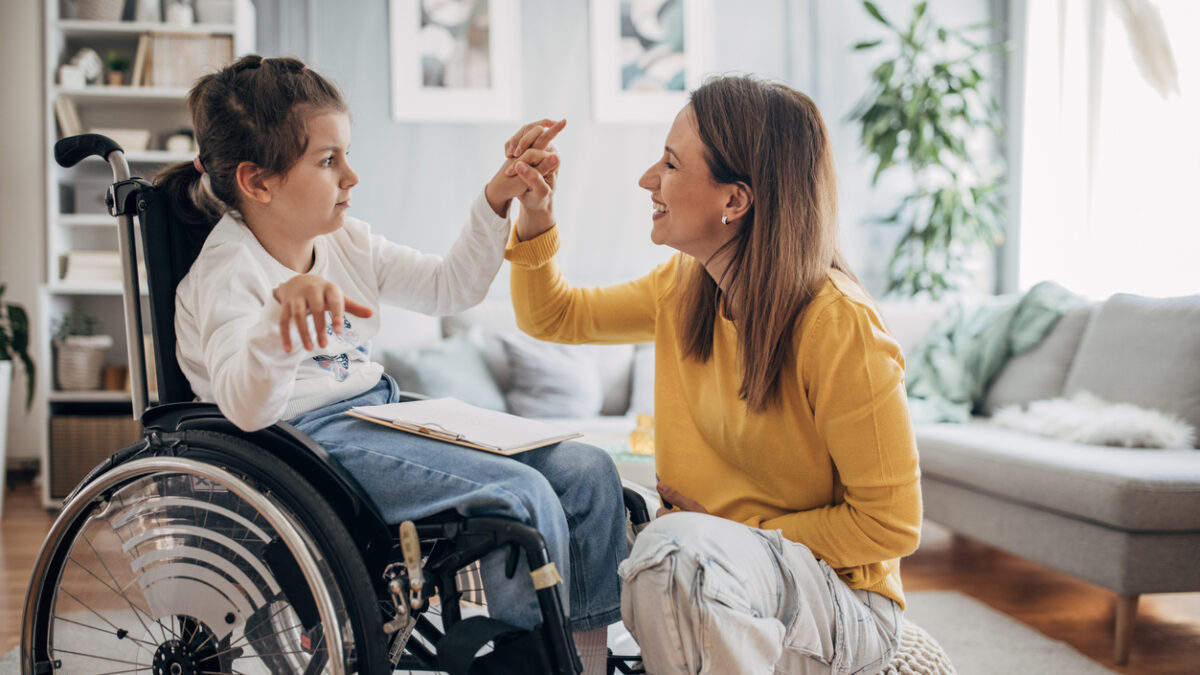 Mother and young daughter who is in wheelchair enjoying together in living room at home.