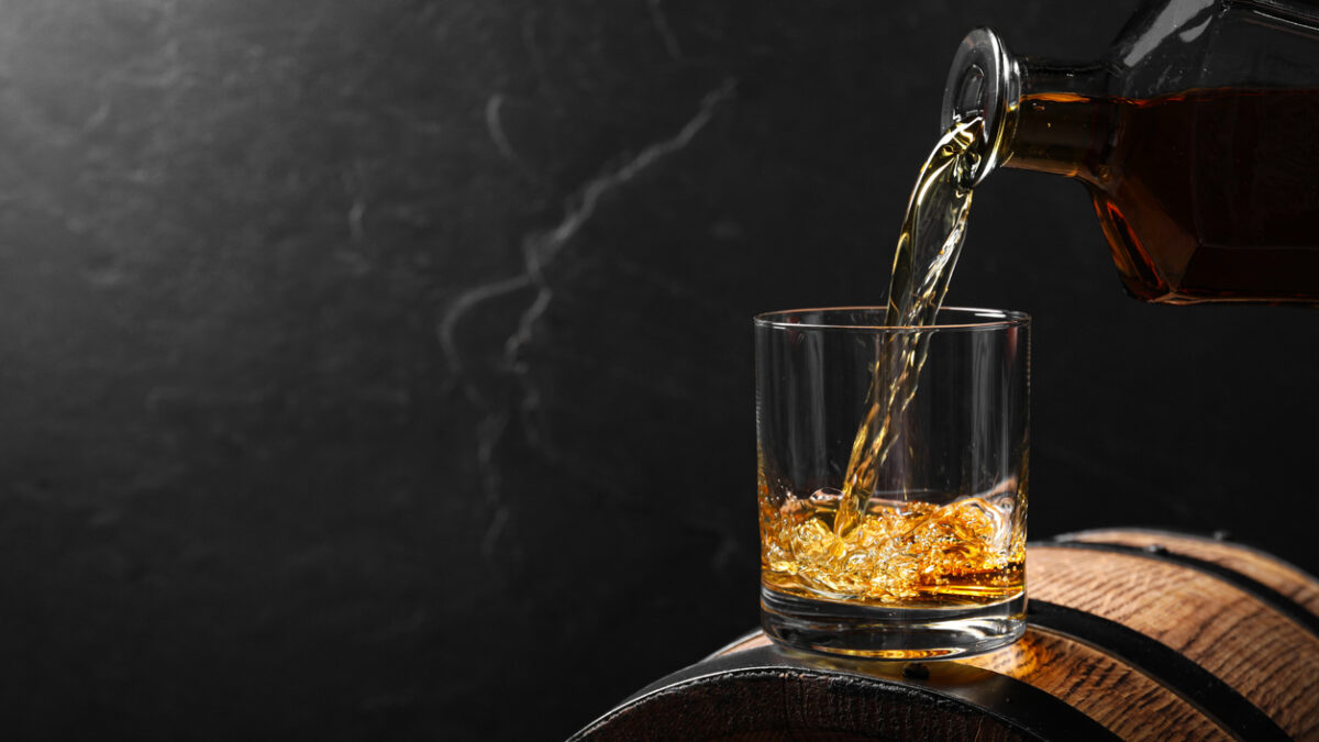 Pouring whiskey into glass from bottle on wooden barrel against grey background, space for text