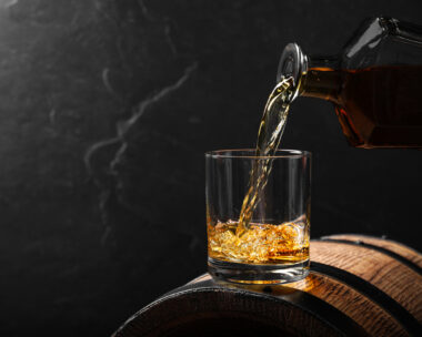 Pouring whiskey into glass from bottle on wooden barrel against grey background, space for text