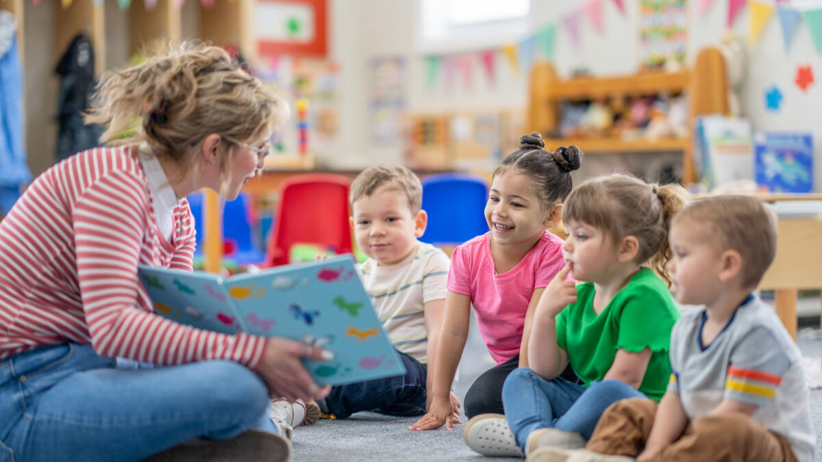A preschool teacher sits on the floor of her classroom with a small group of students as she reads them a book. The children are each dressed casually and are focused on the story.