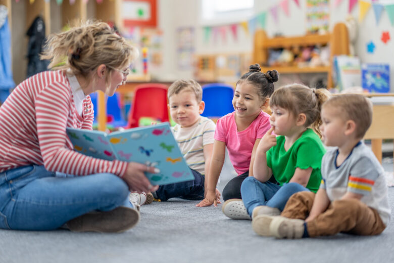 A preschool teacher sits on the floor of her classroom with a small group of students as she reads them a book. The children are each dressed casually and are focused on the story.