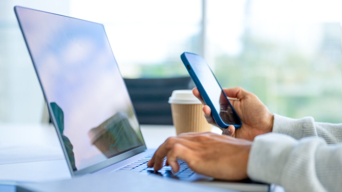 Close up of a Businessman working on a laptop computer and holding and looking at a mobile phone in the office. There are notepads on the desk. He is casually dressed
