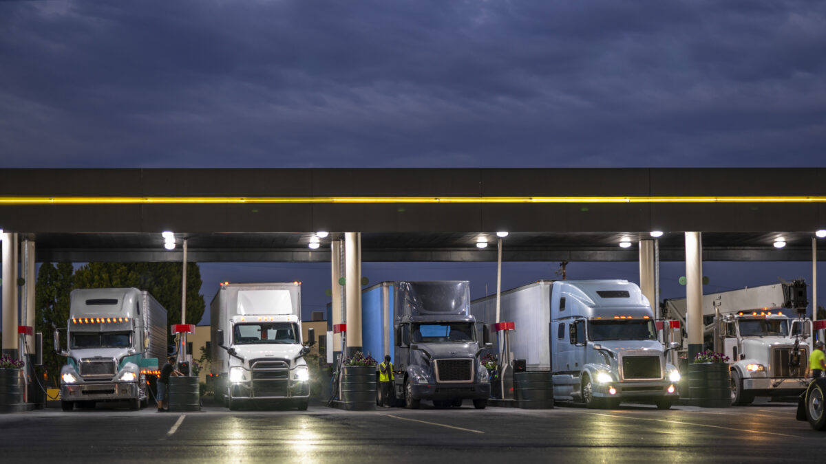 Industrial carriers big rig semi trucks with semi trailers fill the tanks with diesel fuel standing on the truck stop gas station with lights at night to be ready to continue the route by morning