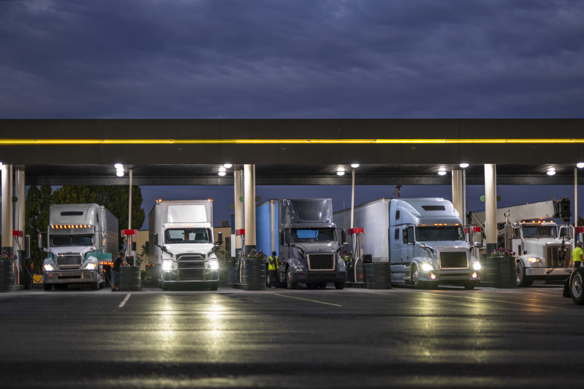 Industrial carriers big rig semi trucks with semi trailers fill the tanks with diesel fuel standing on the truck stop gas station with lights at night to be ready to continue the route by morning