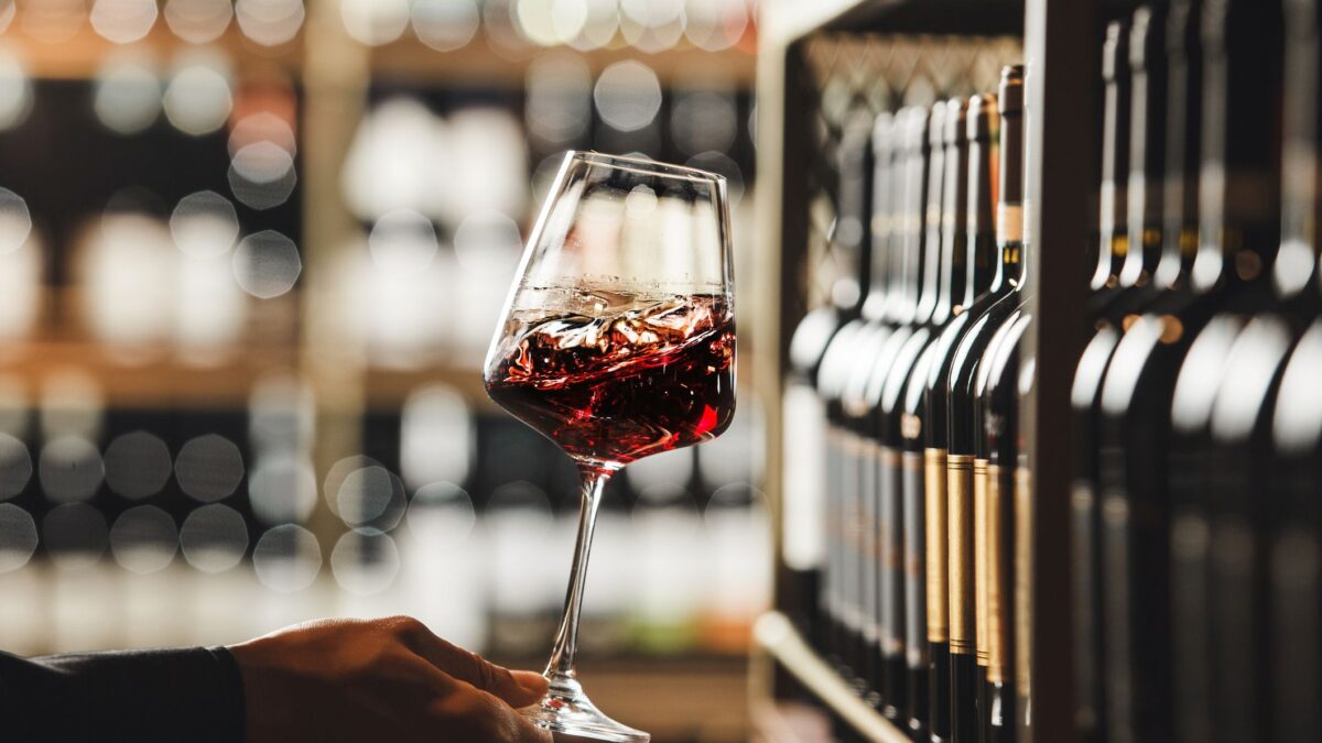 Close up shot of person hand as they swirl a glass of red wine, demonstrating the technique used to aerate the wine and release its aromas and exploring the depth of the wines flavor profile on blurred wine cellar background.
