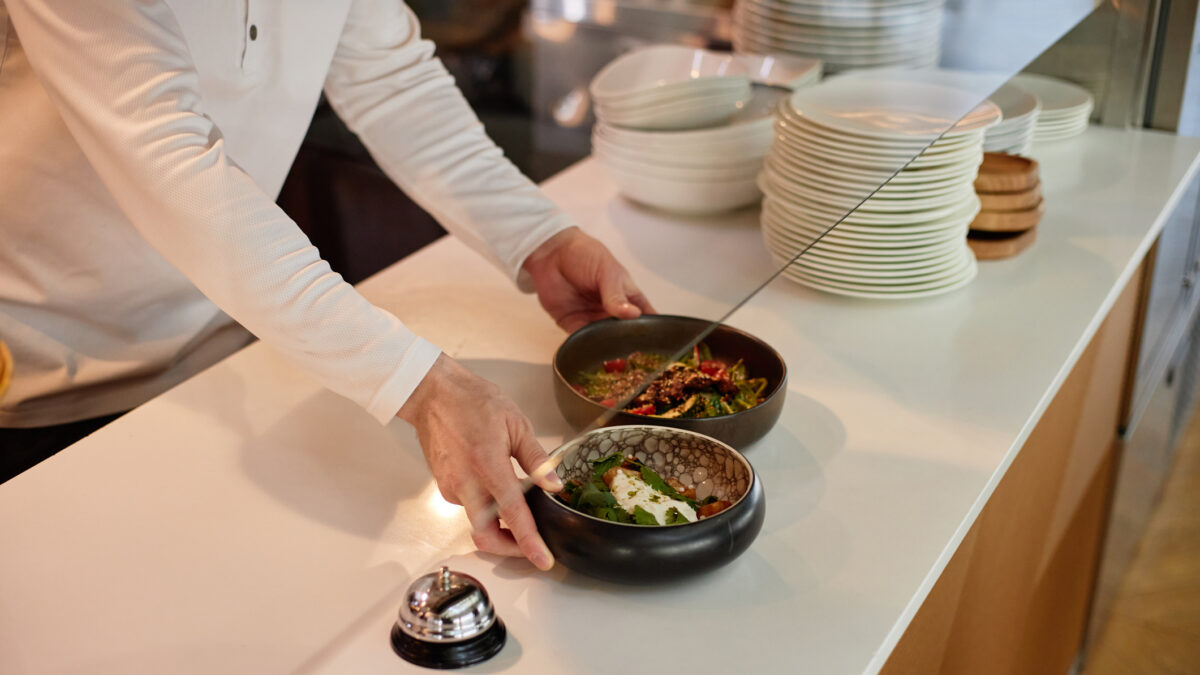 Man placing prepared dishes on counter in restaurant kitchen, hands visible arranging food near service bell, stacks of plates in background