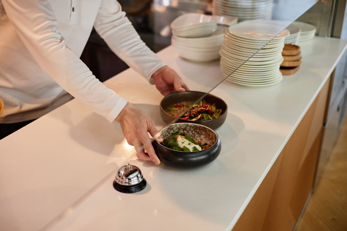 Man placing prepared dishes on counter in restaurant kitchen, hands visible arranging food near service bell, stacks of plates in background