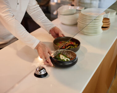 Man placing prepared dishes on counter in restaurant kitchen, hands visible arranging food near service bell, stacks of plates in background