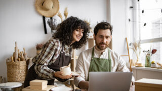 Caucasian couple of co-workers work on small pottery production business. Mature man and young curly-haired woman in aprons use laptop to take inventory of products on store website.