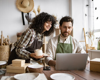 Caucasian couple of co-workers work on small pottery production business. Mature man and young curly-haired woman in aprons use laptop to take inventory of products on store website.
