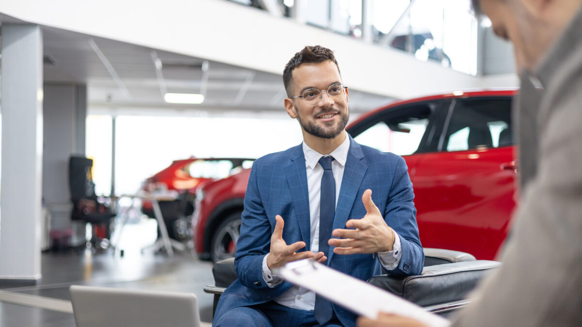 A car dealer in a suit is shown talking to a customer inside a car showroom.