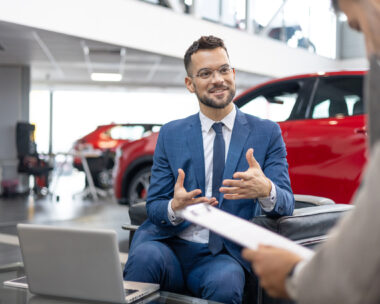 A car dealer in a suit is shown talking to a customer inside a car showroom.