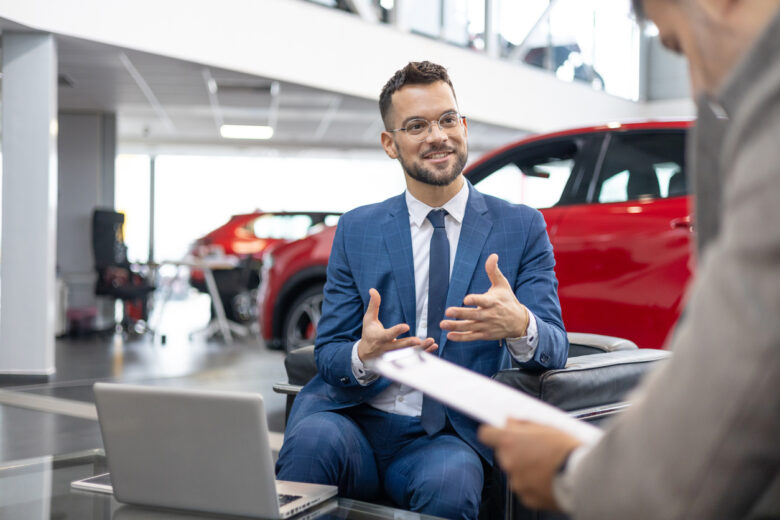 A car dealer in a suit is shown talking to a customer inside a car showroom.