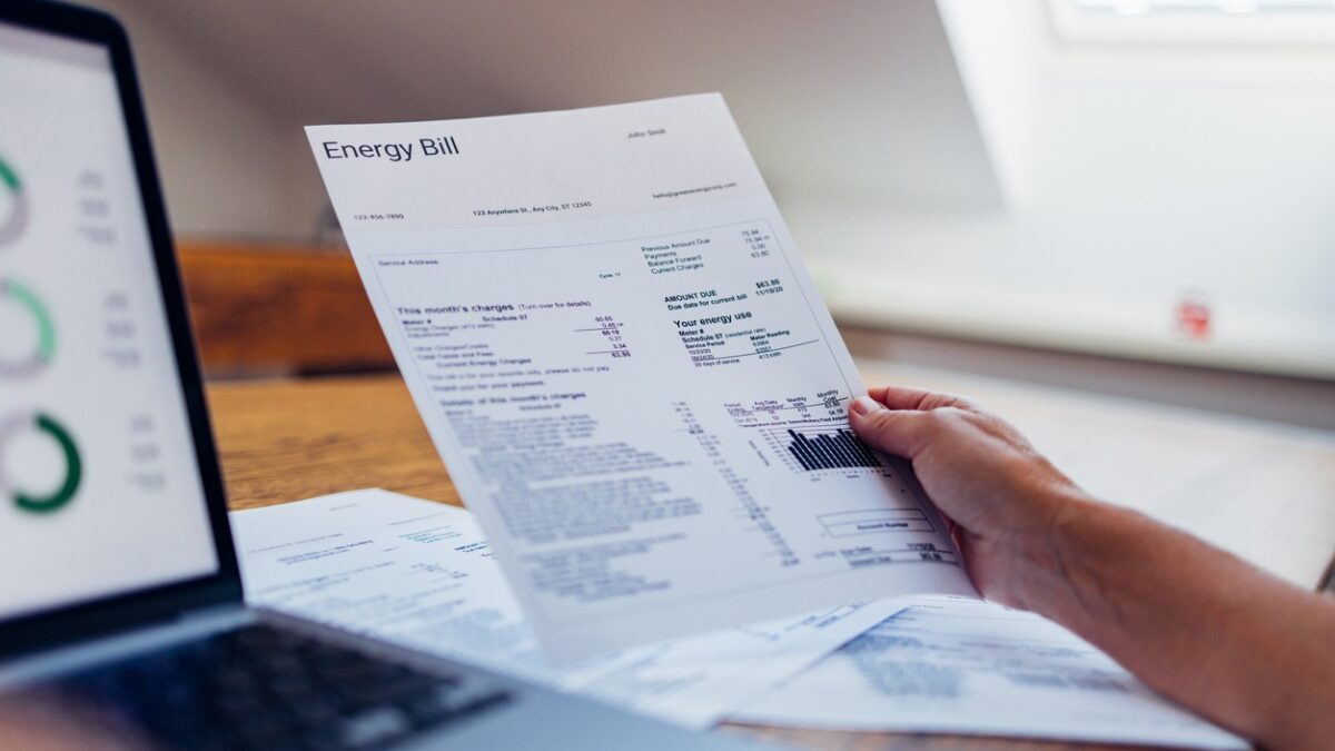 A person reviews their energy bill while using a laptop at home. The document includes various charges and electricity usage information