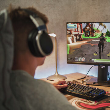 Caucasian teenager wearing headphones plays online video game on PC, spends leisure time at home. Professional young gamer enjoys gameplay in 3D third person shooter on computer. Gaming and Esports