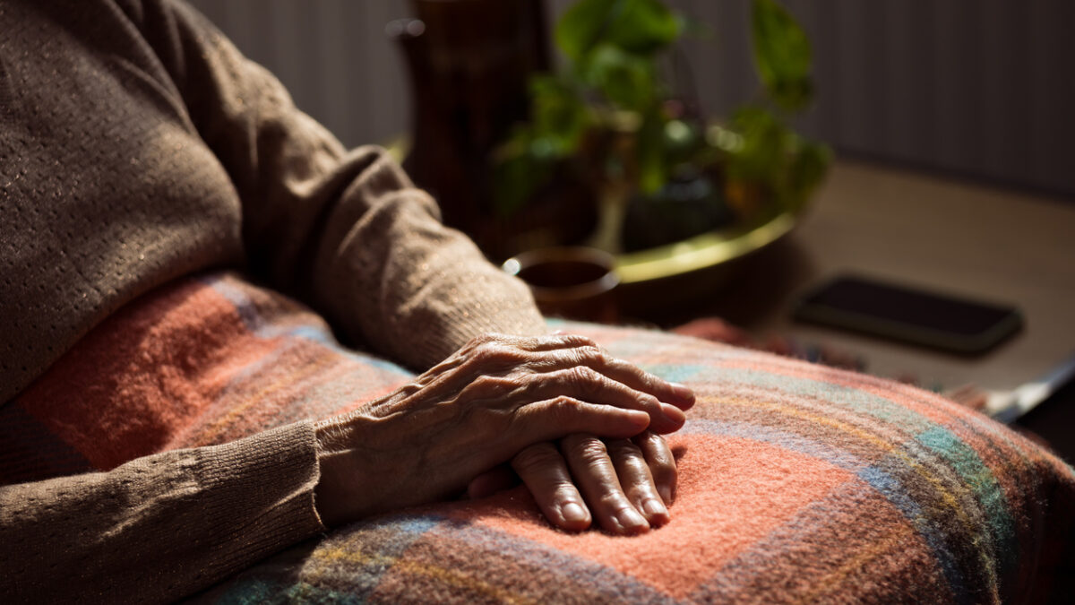 Senior woman sitting in armchair at home with legs covered by blanket. Close up of hands, unrecognizable person.