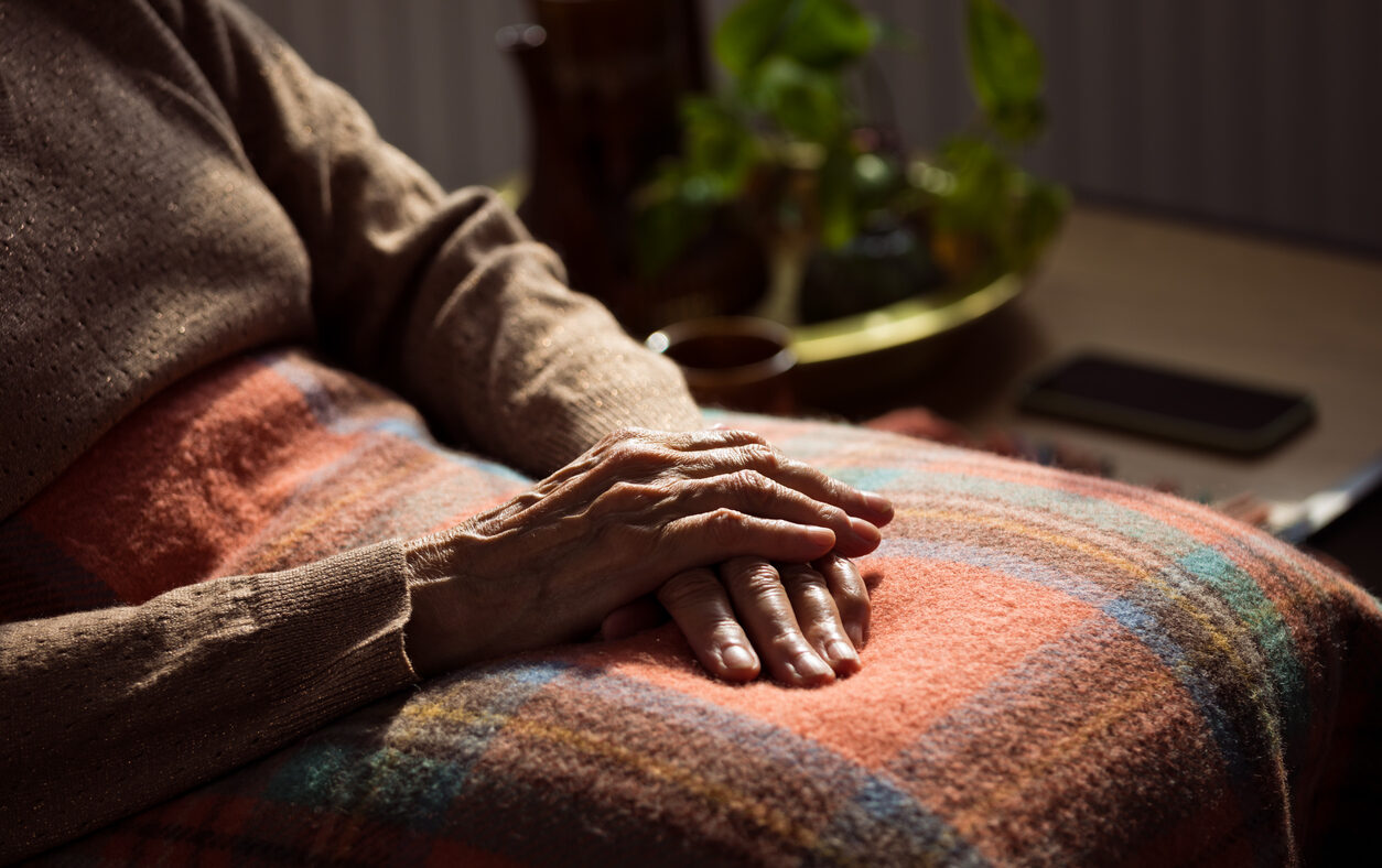 Senior woman sitting in armchair at home with legs covered by blanket. Close up of hands, unrecognizable person.