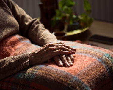 Senior woman sitting in armchair at home with legs covered by blanket. Close up of hands, unrecognizable person.