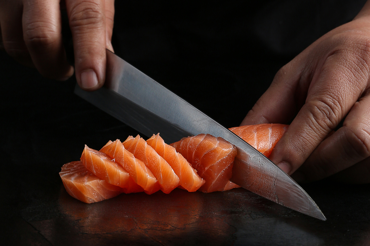 A chef slicing fresh salmon fillet with a sharp Japanese knife, showcasing precision and technique on a dark background.