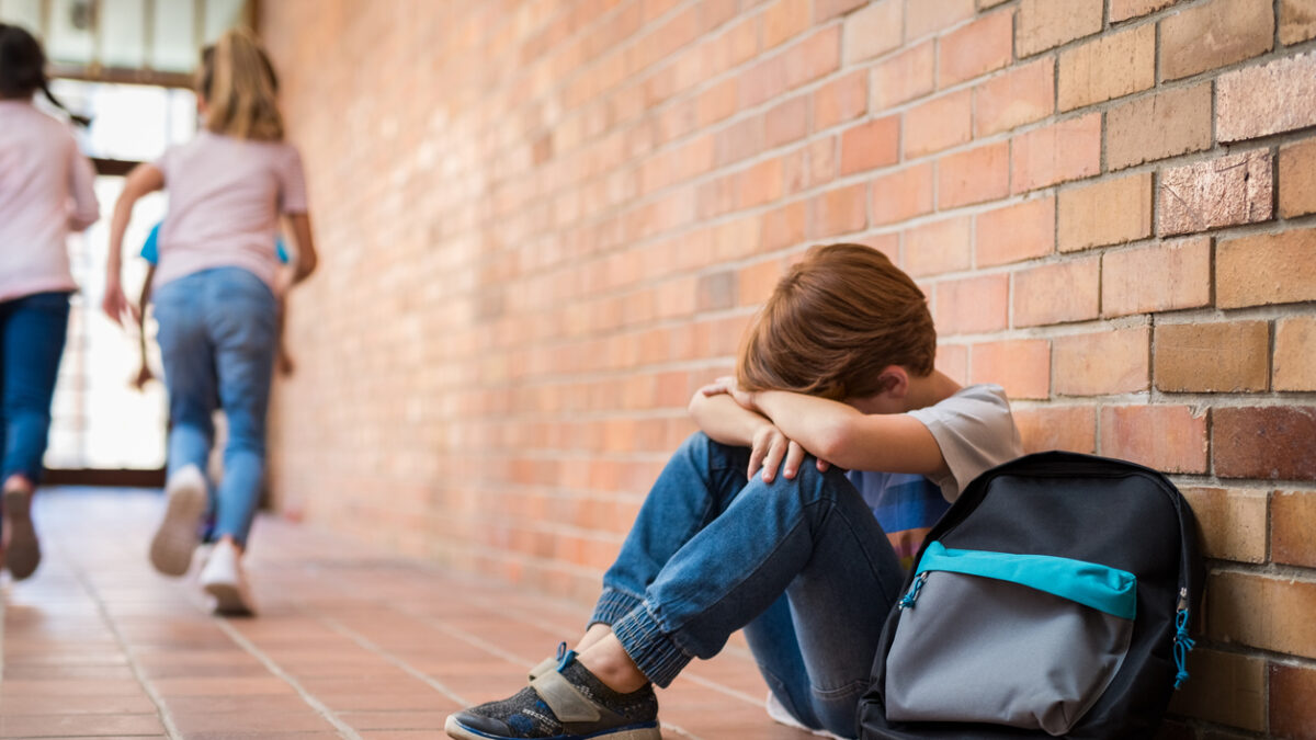Little boy sitting alone on floor after suffering an act of bullying while children run in the background. Sad young schoolboy sitting on corridor with hands on knees and head between his legs.