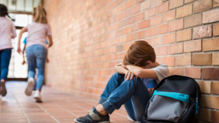 Little boy sitting alone on floor after suffering an act of bullying while children run in the background. Sad young schoolboy sitting on corridor with hands on knees and head between his legs.