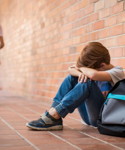 Little boy sitting alone on floor after suffering an act of bullying while children run in the background. Sad young schoolboy sitting on corridor with hands on knees and head between his legs.