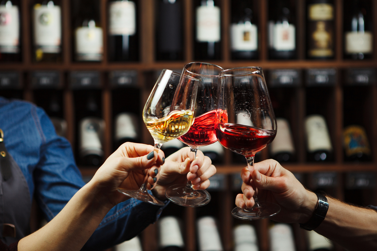 People in good mood clink glasses, celebrating together, photo focus on hands woman and man tasting degustation different types of wine, alcohol beverage to relax, bottles on shelves on background
