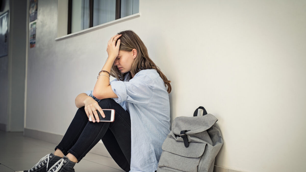 Upset and depressed girl holding smartphone sitting on college campus floor holding head. University sad student suffering from depression sitting on floor at high school. Lonely bullied teen in difficulty with copy space.