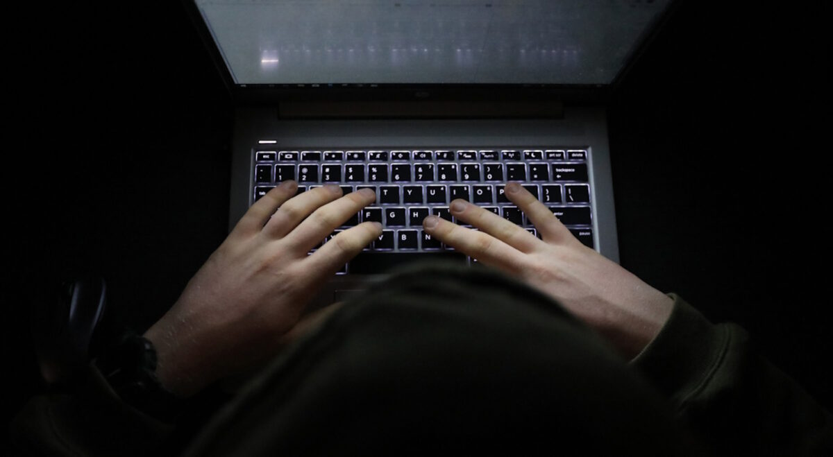 A simple, very dark night time image of hands on an illuminated keyboard typing. Shady person wearing a hood at a computer or laptop in the dark.