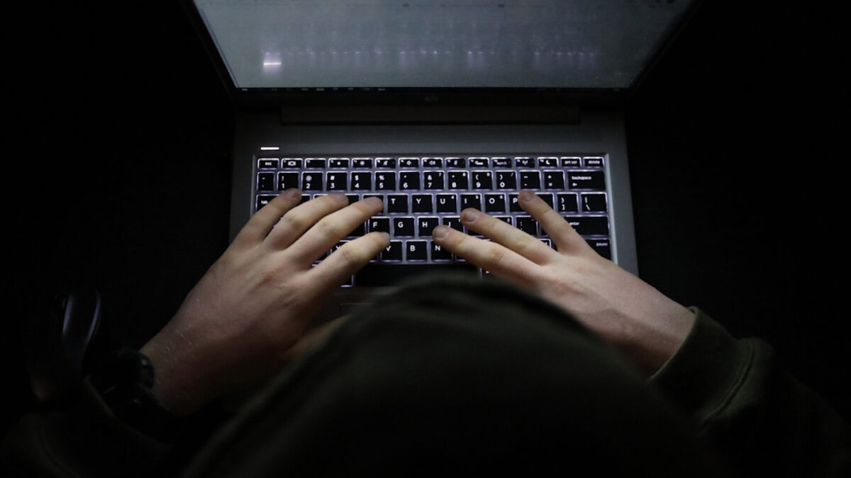 A simple, very dark night time image of hands on an illuminated keyboard typing. Shady person wearing a hood at a computer or laptop in the dark.