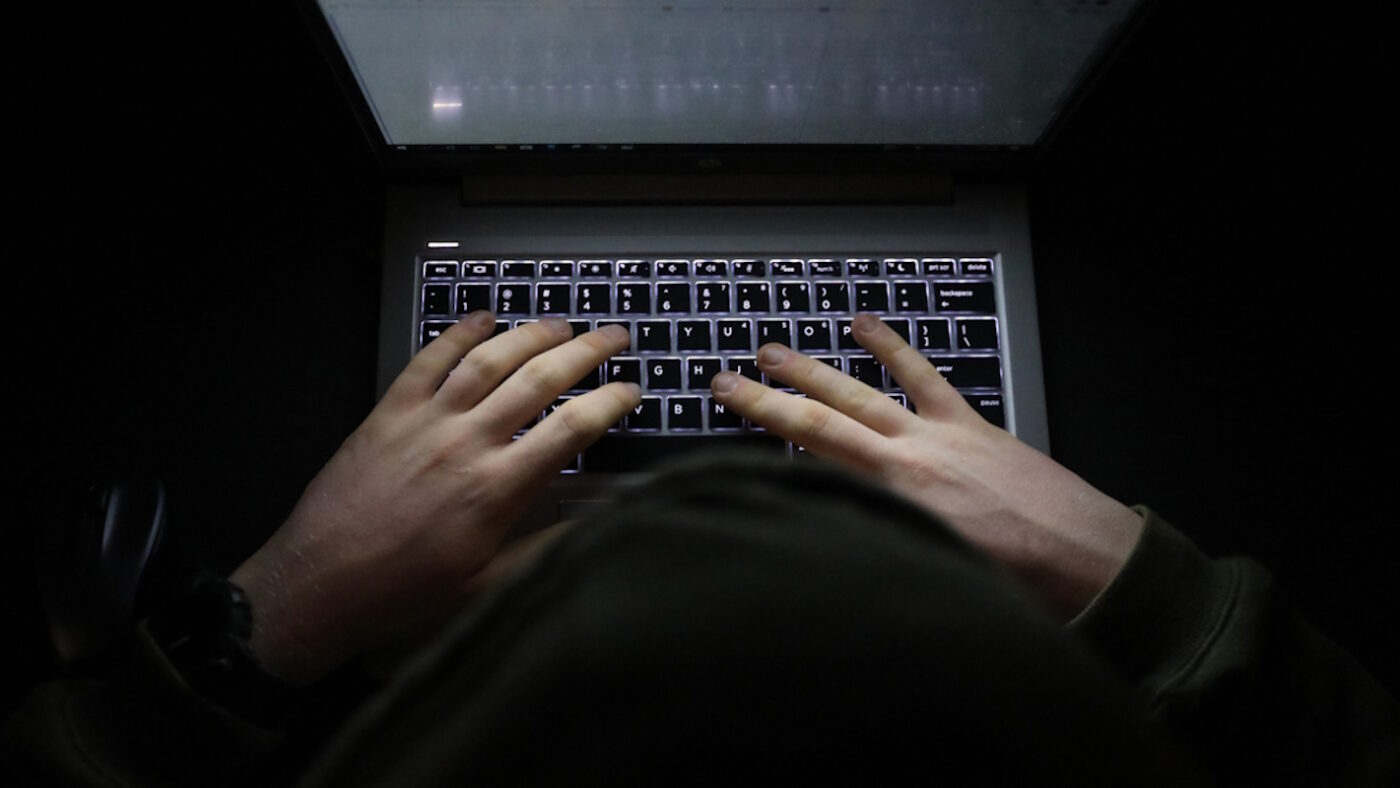 A simple, very dark night time image of hands on an illuminated keyboard typing. Shady person wearing a hood at a computer or laptop in the dark.