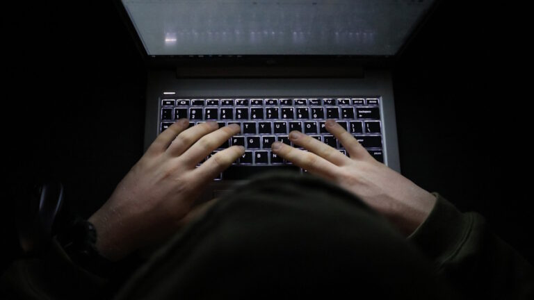 A simple, very dark night time image of hands on an illuminated keyboard typing. Shady person wearing a hood at a computer or laptop in the dark.