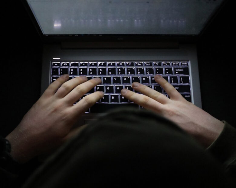 A simple, very dark night time image of hands on an illuminated keyboard typing. Shady person wearing a hood at a computer or laptop in the dark.