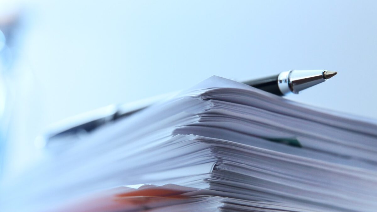 A ballpoint pen rests on top of a stack of documents ready for signing. The image is photographed using a very shallow depth of field with the focus being on the tip of the pen.