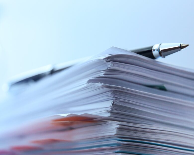 A ballpoint pen rests on top of a stack of documents ready for signing. The image is photographed using a very shallow depth of field with the focus being on the tip of the pen.