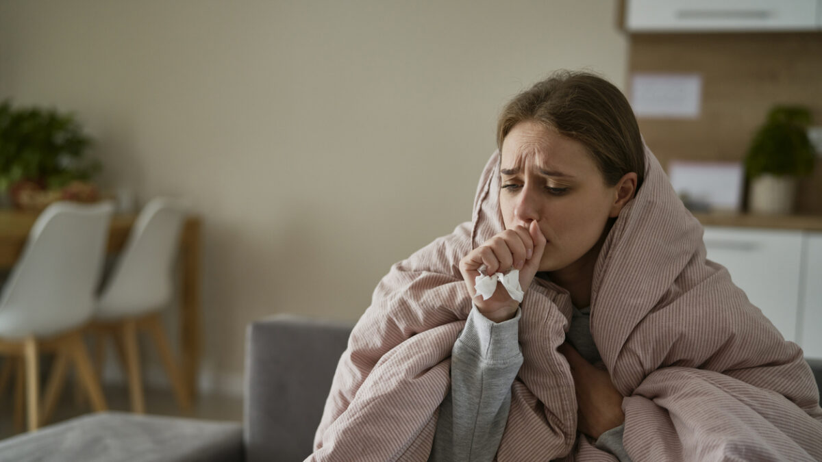 Caucasian young woman coughing and sitting under the duvet at home
