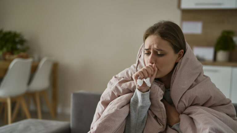 Caucasian young woman coughing and sitting under the duvet at home