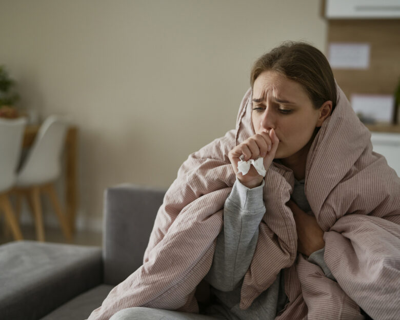 Caucasian young woman coughing and sitting under the duvet at home