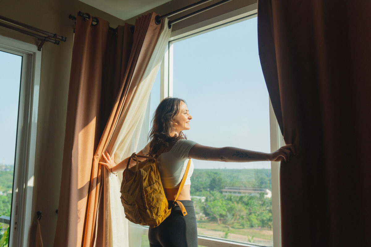 Young woman with backpack walking in to her room in hotel and looking through window on the view of jungles