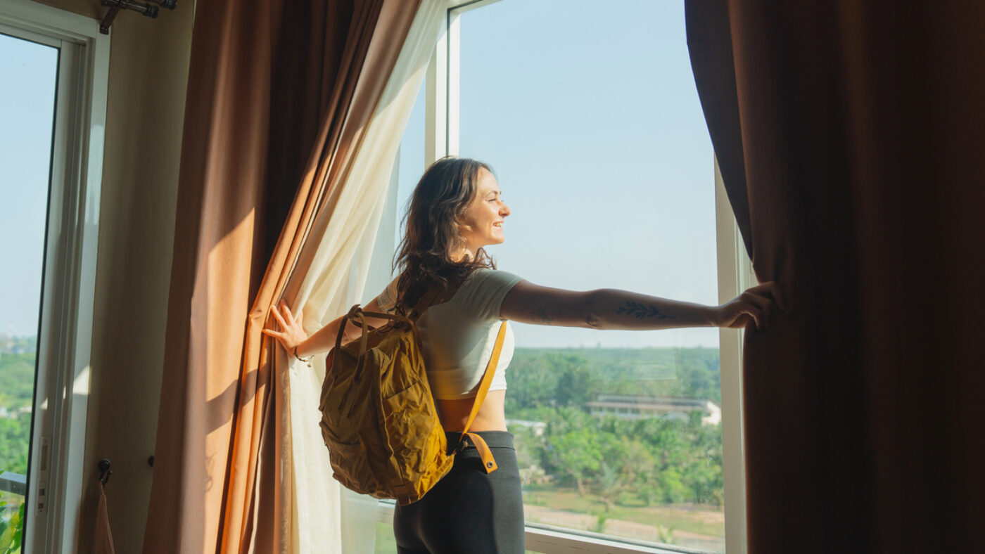 Young woman with backpack walking in to her room in hotel and looking through window on the view of jungles